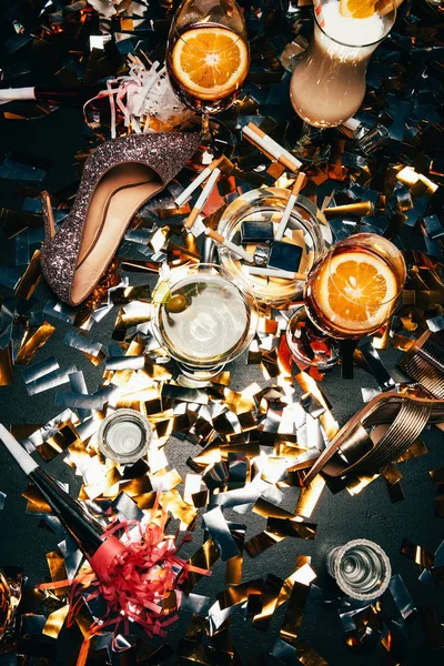 Elevated view of female shoes on high heel, ashtray with cigarettes, alcoholic cocktails and party horns on table covered by golden confetti — Stock Photo