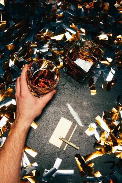 Vista parziale dell'uomo con bicchiere di whisky vicino alla cocaina con banconota arrotolata e carta di credito sul tavolo coperto da confetti dorati — Foto stock