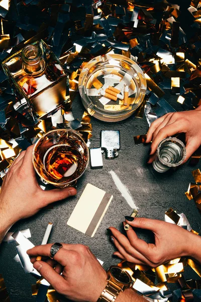 Imagen recortada de pareja celebrando con cocaína, alcohol y cigarrillos en la mesa cubierta por confeti dorado - foto de stock