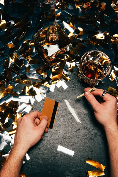 Vista parcial del hombre con billete laminado y tarjeta de crédito que va a oler cocaína en la mesa cubierta por confeti dorado - foto de stock