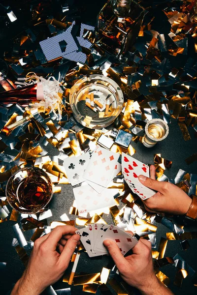Обрезанный образ друзей-мужчин и играющих в покер с алкоголем и сигаретами за столом, покрытым золотыми конфетти — стоковое фото