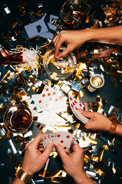 Partial view of men smoking cigarettes and playing poker at table covered by golden confetti — Stock Photo