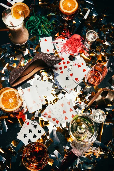 Top view of female shoes on high heels, playing cards, alcoholic cocktails and party horns on table covered by golden confetti — Stock Photo