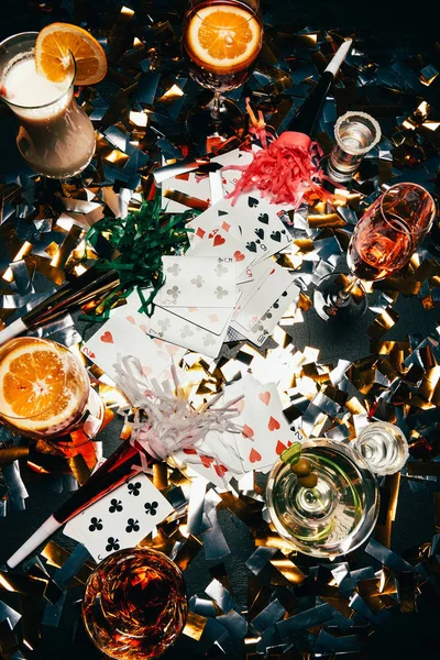 Vista elevada de naipes, cócteles alcohólicos con rebanadas de naranja, whisky y cuernos de fiesta en la mesa cubierta por confeti dorado - foto de stock