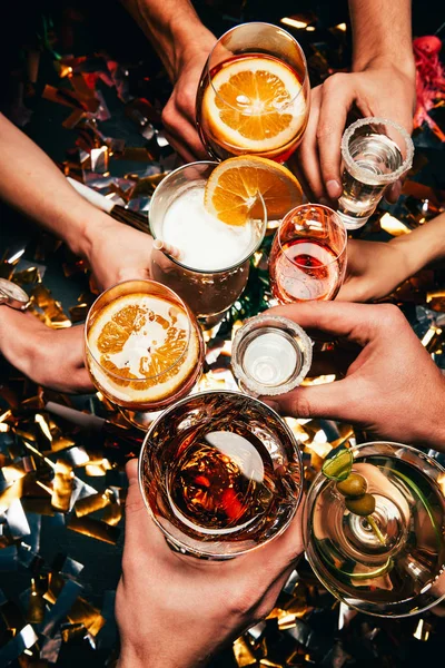 Cropped image of friends clinking by glasses with various alcoholic cocktails at table covered by golden confetti — Stock Photo