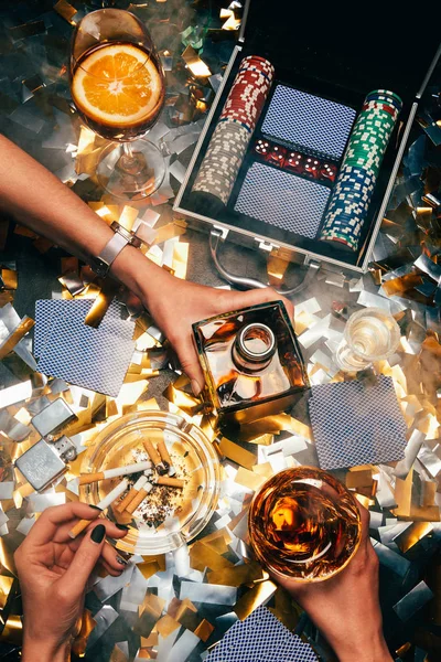 Imagen recortada de mujeres celebrando con alcohol, cigarrillos y fichas de póquer con cartas en la mesa cubierta por confeti dorado - foto de stock