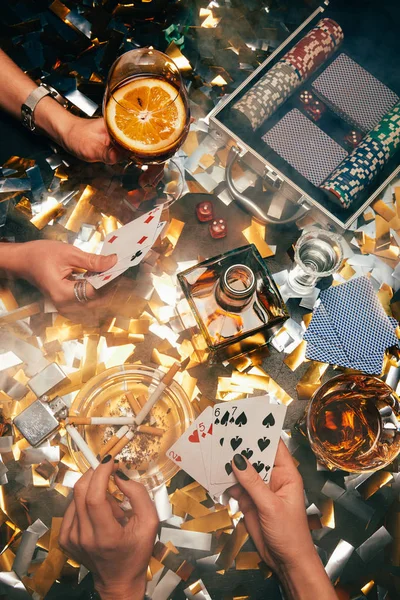 Imagen recortada de las mujeres jugando al póquer en la mesa con whisky, cócteles alcohólicos y cigarrillos y confeti dorado - foto de stock