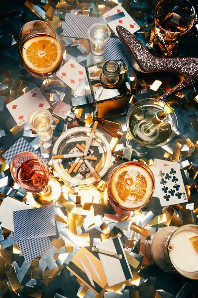 Elevated view of alcoholic cocktails, playing cards, cigarettes, rolled banknote, credit cards and cocaine on table covered by golden confetti — Stock Photo