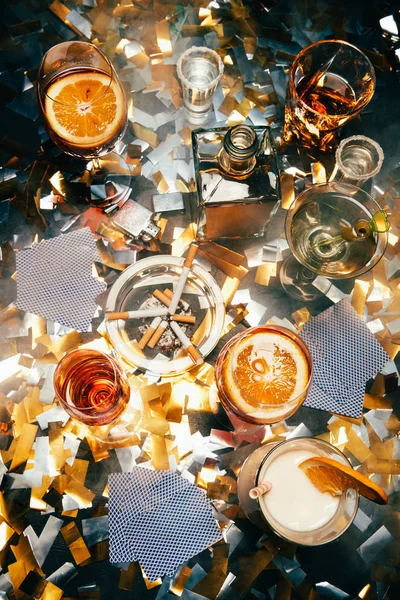 Flat lay com cigarros, coquetéis alcoólicos, uísque e cartas de baralho na mesa cobertas por confetes dourados — Fotografia de Stock