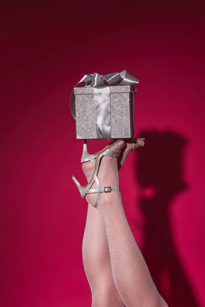 Cropped image of woman in high heels holding silver gift box on legs at party on burgundy — Stock Photo