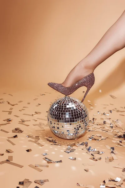 Cropped image of woman putting leg on silver disco ball, confetti on floor — Stock Photo