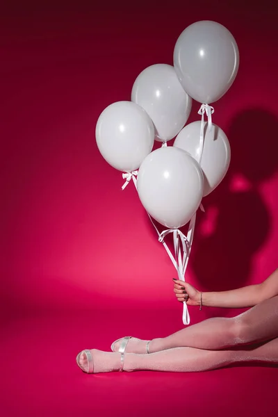 Cropped image of girl sitting and holding bundle of white balloons at party on burgundy — Stock Photo