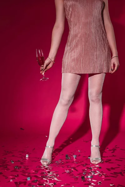 Low section of woman in dress standing on confetti with glass of champagne at party on burgundy — Stock Photo