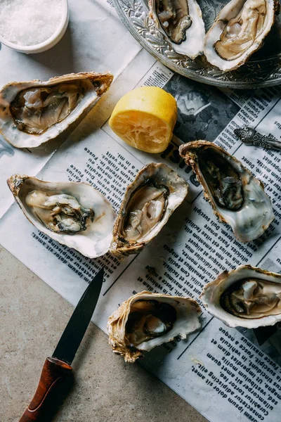 Plat avec huîtres, citron et couteau sur papier journal sur surface grise — Photo de stock