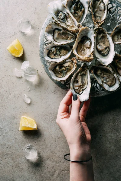 Partial view of woman taking oyster from bowl on grey tabletop with ice and lemon pieces — Stock Photo