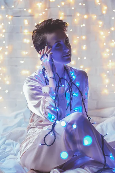 Pensive woman in pajamas with christmas lights resting on bed at home — Stock Photo