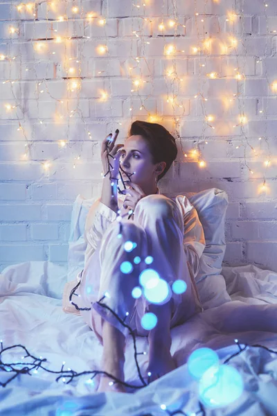 Nachdenkliche Frau im Schlafanzug mit Weihnachtsbeleuchtung, die zu Hause im Bett liegt — Stockfoto