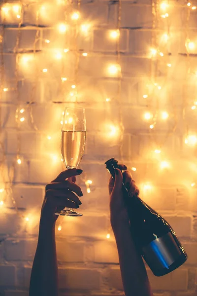 Partial view of woman holding glass and bottle of champagne in hands with christmas lights behind — Stock Photo