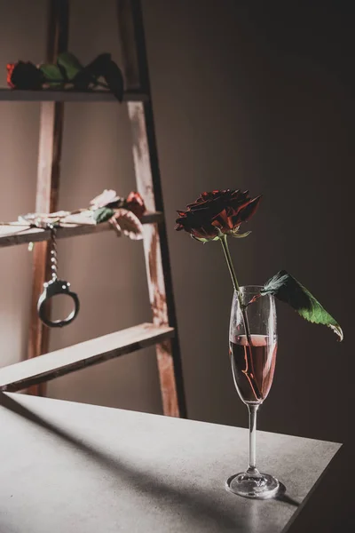 Vermelho rosa flores em taça de champanhe na mesa de pedra com escada de madeira e algemas de metal no fundo — Fotografia de Stock