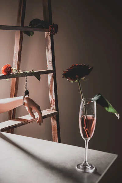 Rote Rosenblüten in Champagnerglas auf Steintisch mit Holzleiter und Frau in Metallhandschellen auf Hintergrund — Stockfoto
