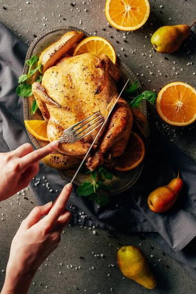 Cropped view of woman cutting fried chicken with oranges, pears and greenery on tray — Stock Photo