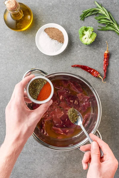 Vue recadrée de la femme ajoutant des épices dans la viande crue en pot avec de l'huile, des poivrons, du brocoli et du romarin — Photo de stock