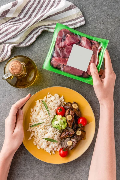 Cropped view of woman holding plate with rice, meat and broccoli on plate with raw meat — Stock Photo