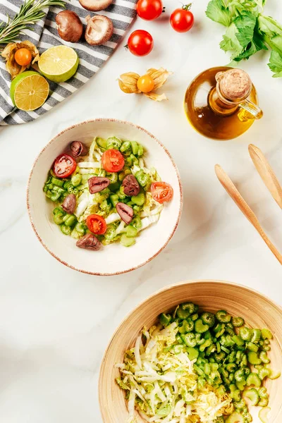 Top view of salad, cabbage and celery in bowls with bottle of oil, cherry tomatoes, limes and mushrooms — Stock Photo