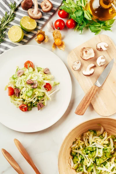 Ensalada, col y apio en plato con botella de aceite, tomates cherry, limas y champiñones en tabla de cortar - foto de stock