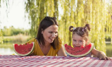 Anne ve kızı nehir yakınındaki parkta bir karpuz dilimi zevk. Anne ve kızı dışarıda birlikte vakit geçirirken meyve keyfi. Ön görünüm.