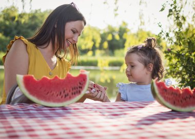 Bekar bir anne ve kızı masada oturup karpuz dilimi yiyor. Kızlar masada oturup karpuz dilimi yiyorlar. Tatlı meyveden sonra peçeteyle kızını temizleyen bir anne. Meyve tüketen