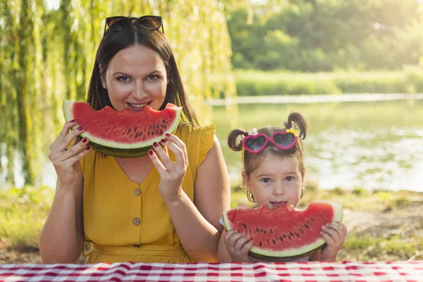 Bekar bir anne ve kızı masada oturup karpuz dilimi yemeye hazırlanıyorlar. Anne ve kızı ellerinde karpuz dilimi ni tutup kameraya bakıyorlar. Ön görünüm.