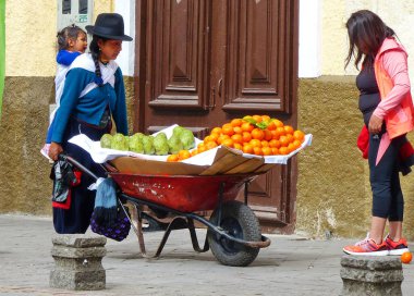 Cuenca, Ekvador - 27 Ekim 2018: Genç kadın sokak satıcısı sırtında çocuk taşıyor ve meyve Cherimoya ve mandalina satıyor. Ekvador 'da meyve satmak adettendir.