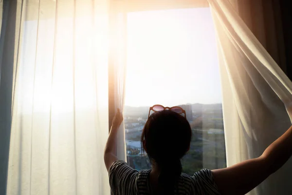 Woman is opening the curtain at the window in the morning.