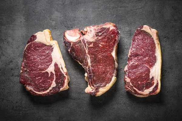 Yaşlı sığır eti biftek - antrikot, striploin, t-bone biftek siyah arduvaz arka plan üzerine kuru. Kopya alanı ile üstten görünüm.