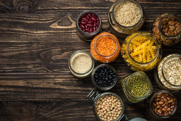 Cereales, legumbres y frijoles en frascos de vidrio sobre mesa de madera oscura . — Foto de Stock