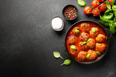 Köfteler domates sosunda, demir tabakta, siyah zemin üzerinde, yemek pişirmek için malzemelerle. Metin için boşluk olan üst görünüm.