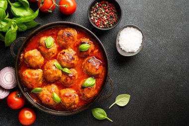Köfteler domates sosunda, demir tabakta, siyah zemin üzerinde, yemek pişirmek için malzemelerle. Metin için boşluk olan üst görünüm.