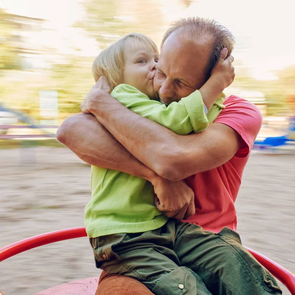 小さな男の子を抱きしめるとカルーセルに乗っている間彼の父のキスします 正方形の比で引き締まったイメージ — ストック写真