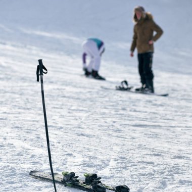 Bir çift kayak ve sopa karda sıkışmış, insanlar arka planda.