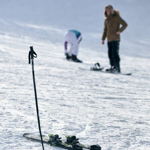 Bir çift kayak ve sopa karda sıkışmış, insanlar arka planda.
