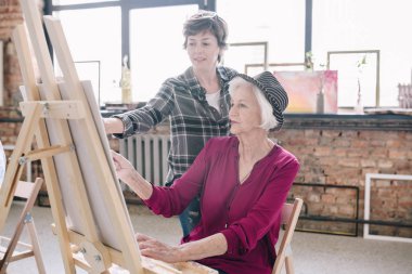 Şövale art Studio, oturmak ve yorum karşı büyük pencereler veren gülümseyen kadın öğretmen ile sanat zevk boyama zarif kadın kıdemli portresi.