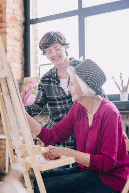 Yan görünümü şövale yorum, güneş ışığı sahnede veren gülümseyen kadın öğretmen ile sanat eğitimi art Studio oturan boyama modern kadın kıdemli portresi