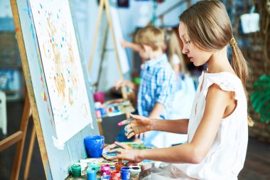Side view portrait of talented teenage girl painting beautiful picture on easel in art class, working with other children clipart