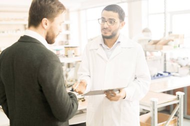 Genç Arap mühendisi giyen laboratuvar kat holding Pano elinde duran modern fabrikasında üretim departmanı, sakallı çift ile başarılı iş sonuçlarını tartışırken.