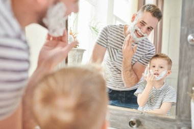 Yakışıklı baba şirin küçük oğlu tıraş kullanmayı öğretmek sıcak tonlu portresi, köpük modern banyo ayna yüzler ile karşı ayakta kaplı