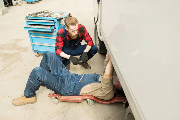 Professional male automobile technician lying on creeper and fixing breakdown of van in service garage, young bearded coworker helping him