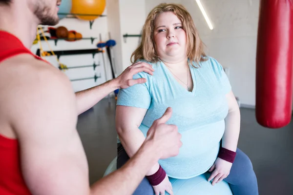 Portrait of obese young woman listening to motivation speech from personal fitness instructor showing thumbs up in gym