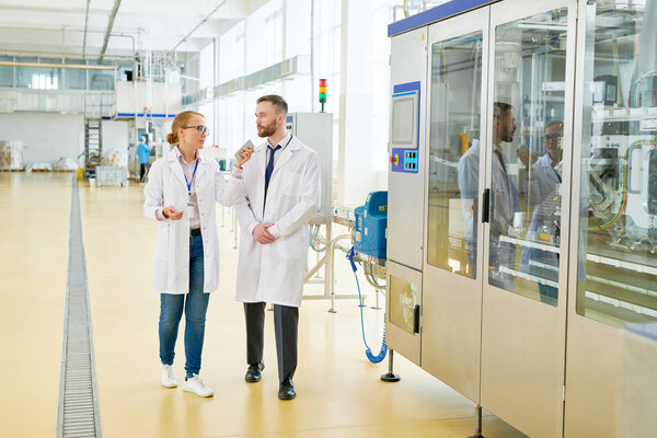 Pretty fair-haired entrepreneur wearing white coat holding smartphone in hand while giving tour around production department of dairy plant for bearded inspector