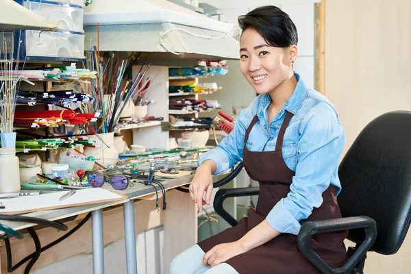 Waist up portrait of smiling Asian woman wearing apron sitting at desk in creative workshop, copy space